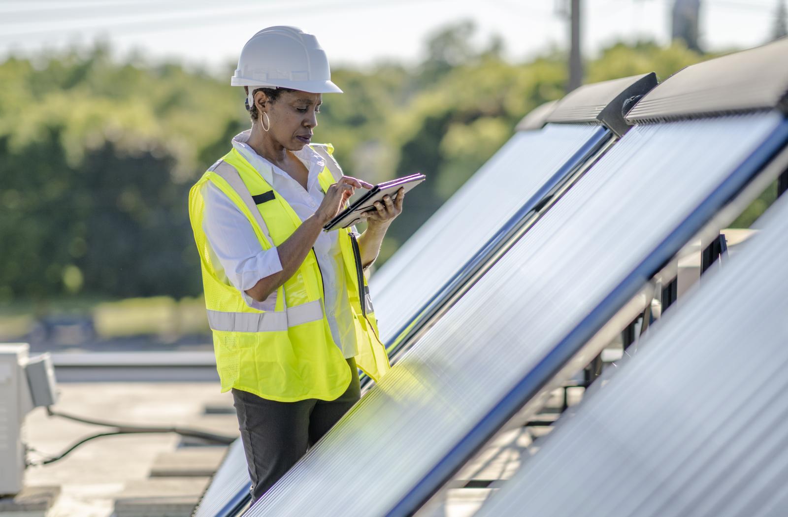 A woman in a hard hat writes in a notebook next to a solar panel